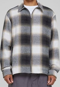 Mężczyzna ubrany w kurtkę z zamkiem błyskawicznym w dużą czarno-białą beżową kratę oraz teksturowany materiał, na tle białej koszuli.
