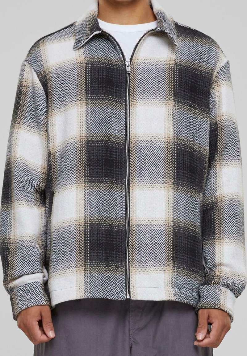 Mężczyzna ubrany w kurtkę z zamkiem błyskawicznym w dużą czarno-białą beżową kratę oraz teksturowany materiał, na tle białej koszuli.