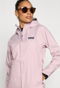 Αδιάβροχο τζάκετ σε ανοικτό ροζ χρώμα με εμπρόσθιο φερμουάρ, κουκούλα και ρυθμιζόμενα κορδόνια. Διαθέτει λογότυπο Patagonia και πλαϊνές τσέπες. Καφέ γυαλιά ηλίου.