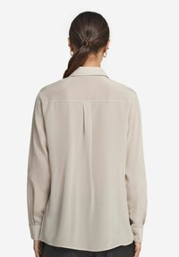 Beige Bluse mit langen Ärmeln und glatter Textur, ausgestattet mit einer Rückenfalte und geknöpften Manschetten. Das einfache Kragendesign unterstreicht ihren klassischen Stil.