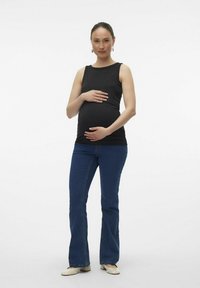 Top negro sin mangas, jeans de campana de mezclilla, zapatos planos de color claro. El sujeto sostiene su barriga de embarazo, de pie sobre un fondo liso.