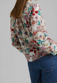 Blouse florale en tissu léger et texturé dans des tons doux de rose, rouge et vert, avec une coupe décontractée et un design d'épaules froncées.