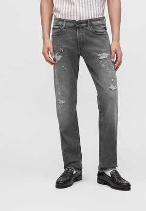 Personne portant un jean gris usé, des mocassins noirs cloutés et une chemise rayée rentrée, debout devant un fond uni.