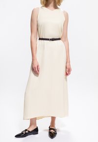 Femme portant une robe maxi sans manches crème avec une ceinture noire et des ballerines noires à bout pointu, se tenant devant un fond blanc.