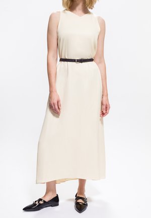 Femme portant une robe maxi sans manches crème avec une ceinture noire et des ballerines noires à bout pointu, se tenant devant un fond blanc.
