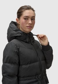 Black puffer jacket with a high collar, zip closure, and drawstring details. Textured fabric and ribbed cuffs enhance warmth.