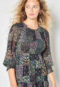 Giovane donna con capelli biondi ricci che indossa un vestito a maniche lunghe con un corpo arricciato e orecchini a cerchio, che guarda di lato.