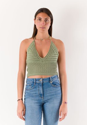 Donna con lunghi capelli scuri che indossa un top verde lavorato a maglia con spalline e jeans blu a vita alta, in piedi davanti a uno sfondo bianco uniforme.