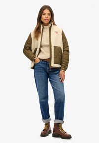 Giubbotto imbottito con davanti in pile beige, maniche verde oliva e due ampie tasche. Indossato sopra un maglione crema a trecce e jeans blu. Stivali marroni con lacci.