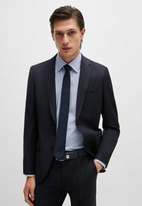 Blazer blu navy con una trama sottile, abbinato a una camicia a righe leggera blu e bianca. Una cravatta e una cintura scure completano il look.