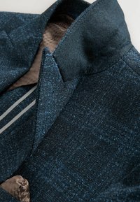 Texturerad mörkblå tyg med en diskret lyster, utrustad med en skarp krage och ett mönstrat brunt foder, kompletterad med kontrasterande vita ränder.