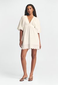 Kurzes weißes Kleid mit Puffärmeln, tiefem V-Ausschnitt, gerafftem Bund und gewelltem Spitzenabschluss. Aus leichtem Stoff mit gestickten Details gefertigt.