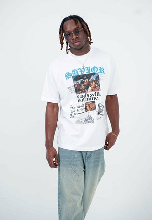 Young man with dreadlocks wearing blue-tinted glasses, a white graphic T-shirt with religious text, and loose blue jeans against a plain background.