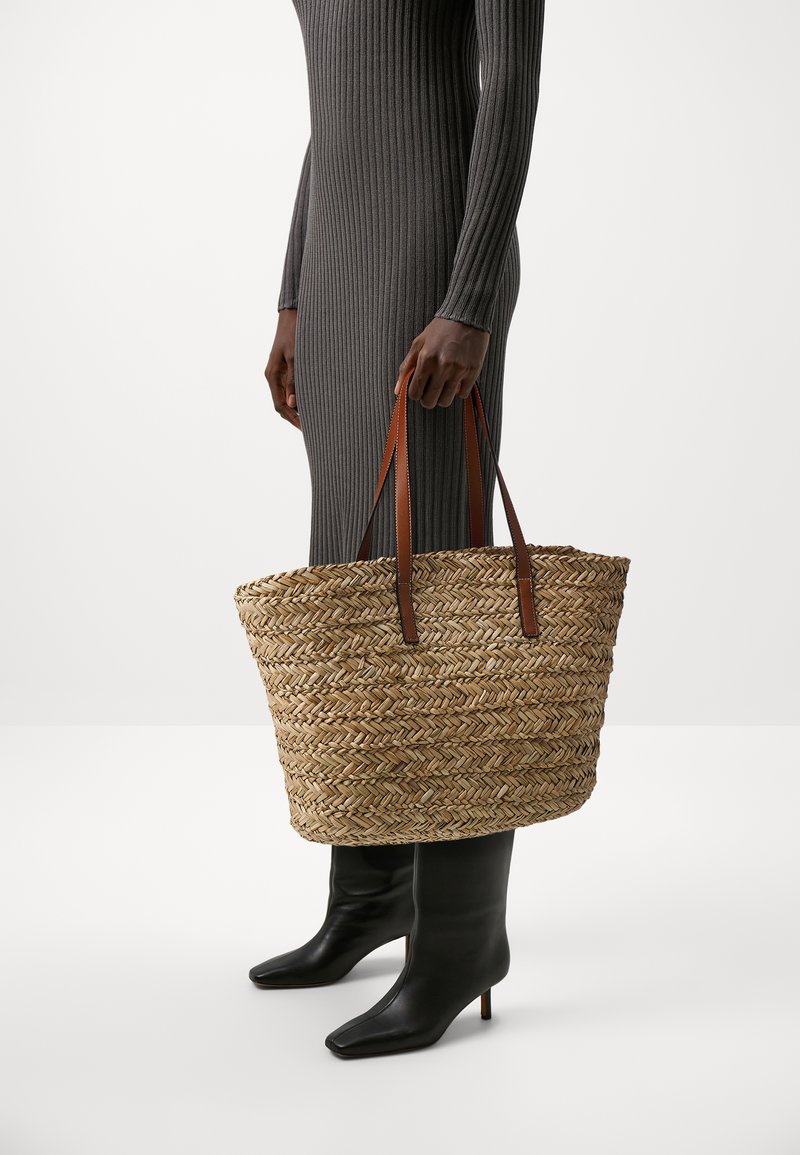 Sac fourre-tout en paille tressée avec des poignées en cuir marron, de forme ovale large, texture détaillée, grande capacité et couleur naturelle claire.