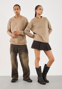 To modeller står mot en hvit bakgrunn iført beige hettegensere; den ene i blek, baggy jeans og sneakers, den andre i et svart skjørt og høye svarte støvler.