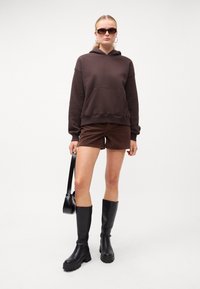 Hanorac maro cu glugă și buzunar frontal, asortat cu pantaloni scurți din catifea maro. Cizme negre până la genunchi și o mică geantă de umăr neagră.