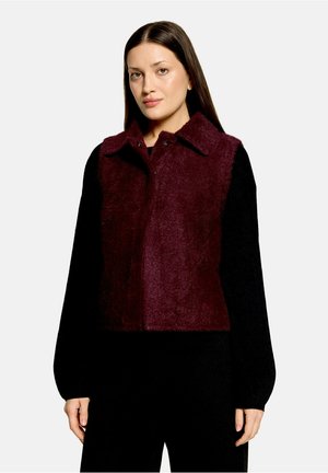 Femme aux cheveux bruns et lisses porte une veste sans manches rouge foncé et duveteuse par-dessus un haut noir à manches longues sur un fond clair uni.