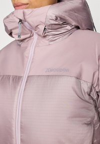 Giacca Houdini di colore rosa pastello con zip frontale, collo alto e parte inferiore testurizzata con linee orizzontali e dettaglio del logo.