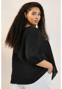 Svart texturerad blus med trekvartsärmar, med en broderad design vid nederkanten och en sidoficka, bärs över ljusa byxor.