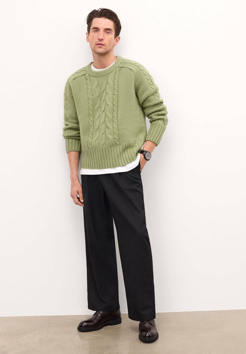 Suéter verde de punto trenzado con dobladillo acanalado, combinado con pantalones negros de corte holgado. Accesorios incluyen un reloj negro y zapatos marrón oscuro.