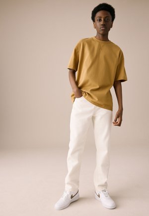 Giovane in piedi con le mani parzialmente nelle tasche, indossa una T-shirt oversize giallo senape, pantaloni bianchi e sneakers bianche con logo beige.