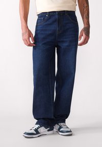 Jean large bleu foncé avec cinq poches, fermeture éclair, et couture contrastante subtile, associés à des baskets vertes et blanches.