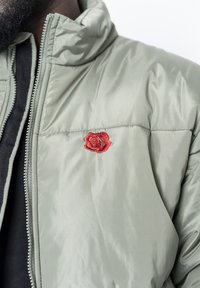 Ljust grön pufferjacka med ståkrage, med en röd ros broderad på bröstet, slät textur och framfickszipp.