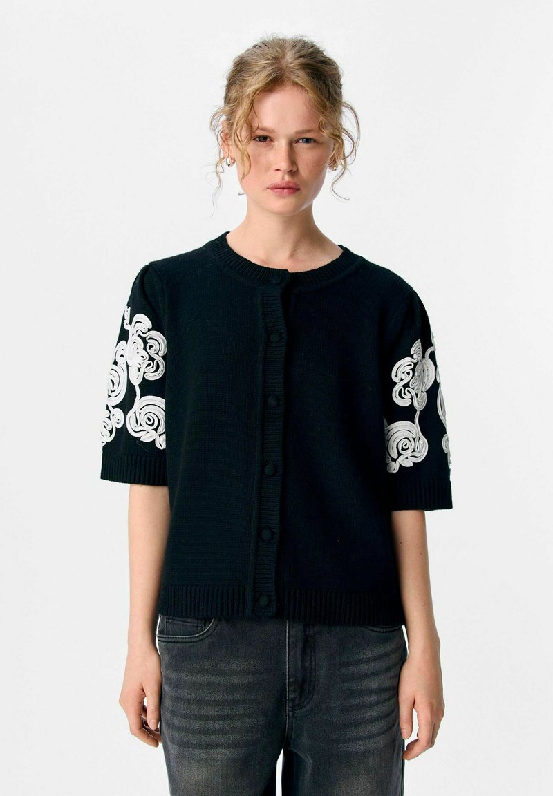 Kvinna med lockigt blont hår bär en svart kortärmad cardigan med vit blommig brodyr på ärmarna, tillsammans med urtvättade svarta jeans.