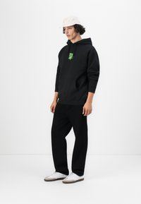 Sort hættejakke med et grønt grafisk logo, frontlomme, båret med løse sorte bukser og hvide sneakers. Baggrunden er hvid.