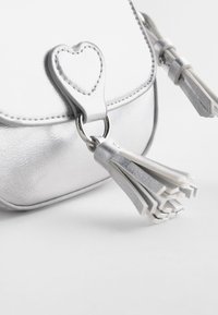 Gros plan sur un coin de sac à main argenté texturé avec un détail en cuir en forme de cœur et deux pampilles en cuir argenté attachées par un anneau métallique.