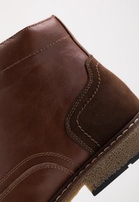Botín de cuero marrón hasta el tobillo con paneles lisos y texturizados, costuras en contraste y una suela de goma duradera con un dibujo en relieve.