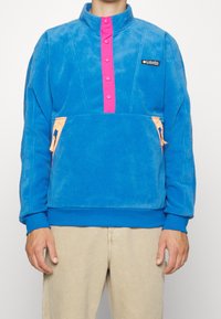 Pullover in pile blu con colletto alto, patta con bottoni rosa, tasche con zip arancioni e polsini a costine. Texture morbida e design sportivo.
