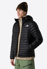 Chaqueta acolchada negra con capucha, con un diseño de acolchado y logo en el pecho. Llevada sobre un forro polar beige y combinada con pantalones verdes.