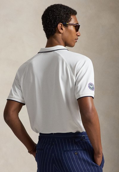 Άνδρας που φορά λευκό πόλο με το λογότυπο του Wimbledon στο μανίκι και navy παντελόνι με λεπτές ρίγες, γυρισμένος με την πλάτη, με τα χέρια στις τσέπες.