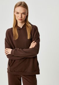Sudadera marrón hecha de un tejido suave y texturizado con un corte relajado, que presenta un bolsillo canguro y una capucha de doble capa.