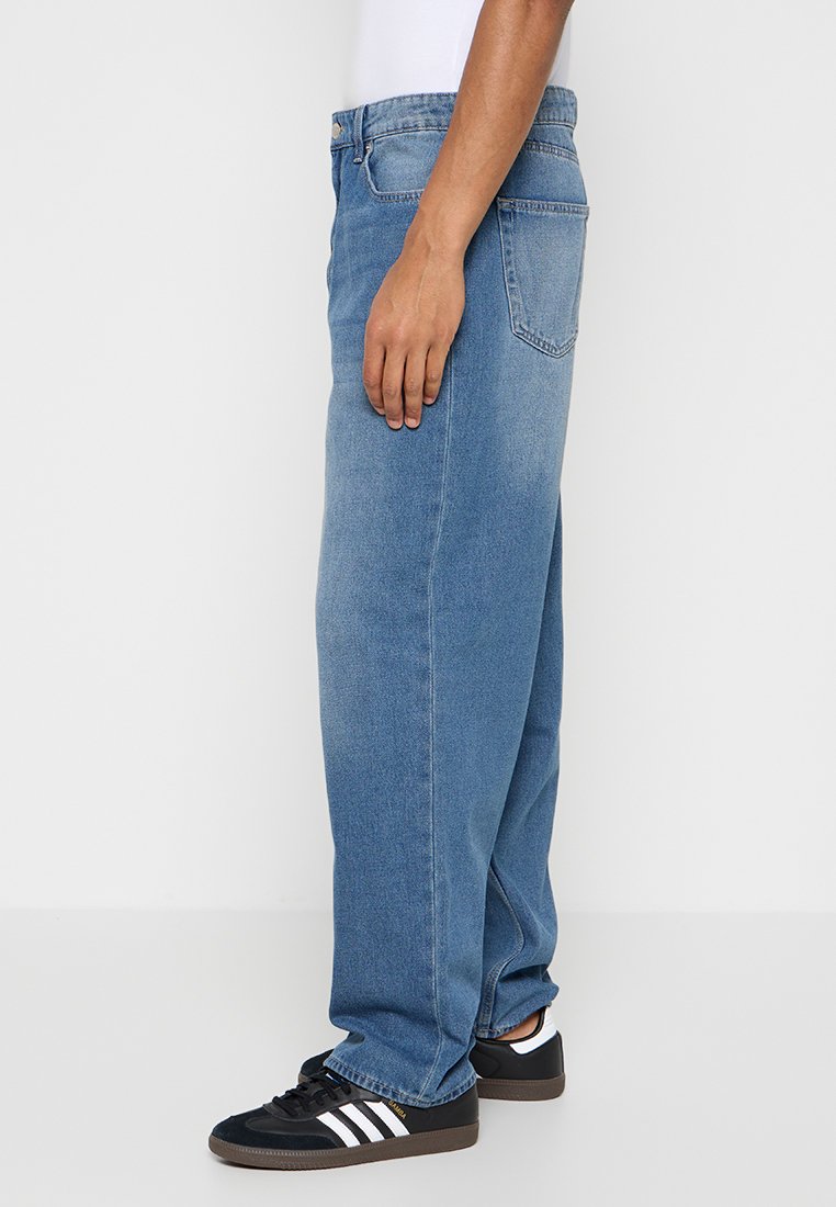 Weit geschnittene blaue Jeans aus Denim mit mittlerer Leibhöhe, Fünf-Taschen-Design, getragen mit schwarzen Sportschuhen mit weißen Akzenten.