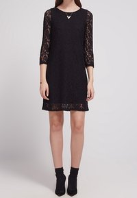 Svart spetsklänning med trekvartsärmar, V-ringning och en nederkant med spetsdetaljer. Bärs med svarta strumpor som går ner till anklarna.