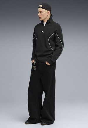 Joven con cabello rubio que lleva un suéter negro de Puma, pantalones anchos negros y una gorra negra puesta al revés, de pie con las manos en los bolsillos.