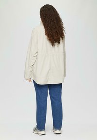 Lichtbeige jas met een losse pasvorm, voorzien van een hoge kraag, elastische taille en lange mouwen, gecombineerd met blauwe jeans en grijze sneakers.
