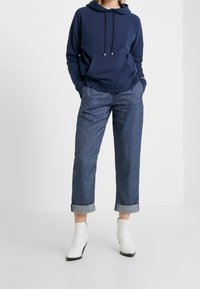Felpa blu navy con tasca frontale e coulisse, abbinata a jeans in denim chiaro arrotolati e stivaletti bianchi alla caviglia.