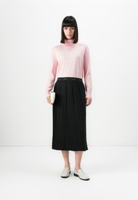 Kobieta w różowym swetrze z golfem, czarnej plisowanej spódnicy midi z paskiem Calvin Klein, białych loafersach i trzymająca małą białą kopertówkę.