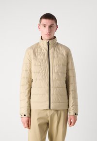 Geacă puffer bej cu model matlasat orizontal, guler înalt, fermoar frontal complet și buzunare laterale. Fabricată dintr-un material neted și ușor.