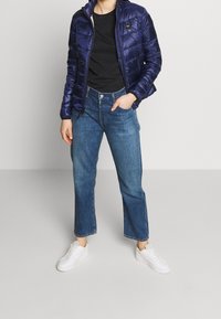 Doudoune marine à fermeture éclair, t-shirt noir, jeans larges bleu clair et baskets blanches, standing contre un fond clair.