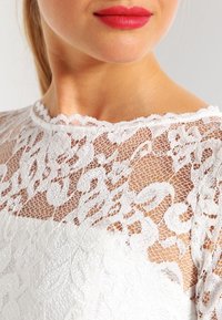 Bílý krajkový top s jemným květinovým vzorem, zvlněným výstřihem a průhlednými detaily. Hladká textura a přiléhavý střih.