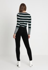 Vrouw die met haar gezicht weg van de camera staat, gekleed in zwarte hoge taille broek, witte haklaarzen tot de enkel en een gestreepte top met lange mouwen met zwarte, witte en groene lijnen.