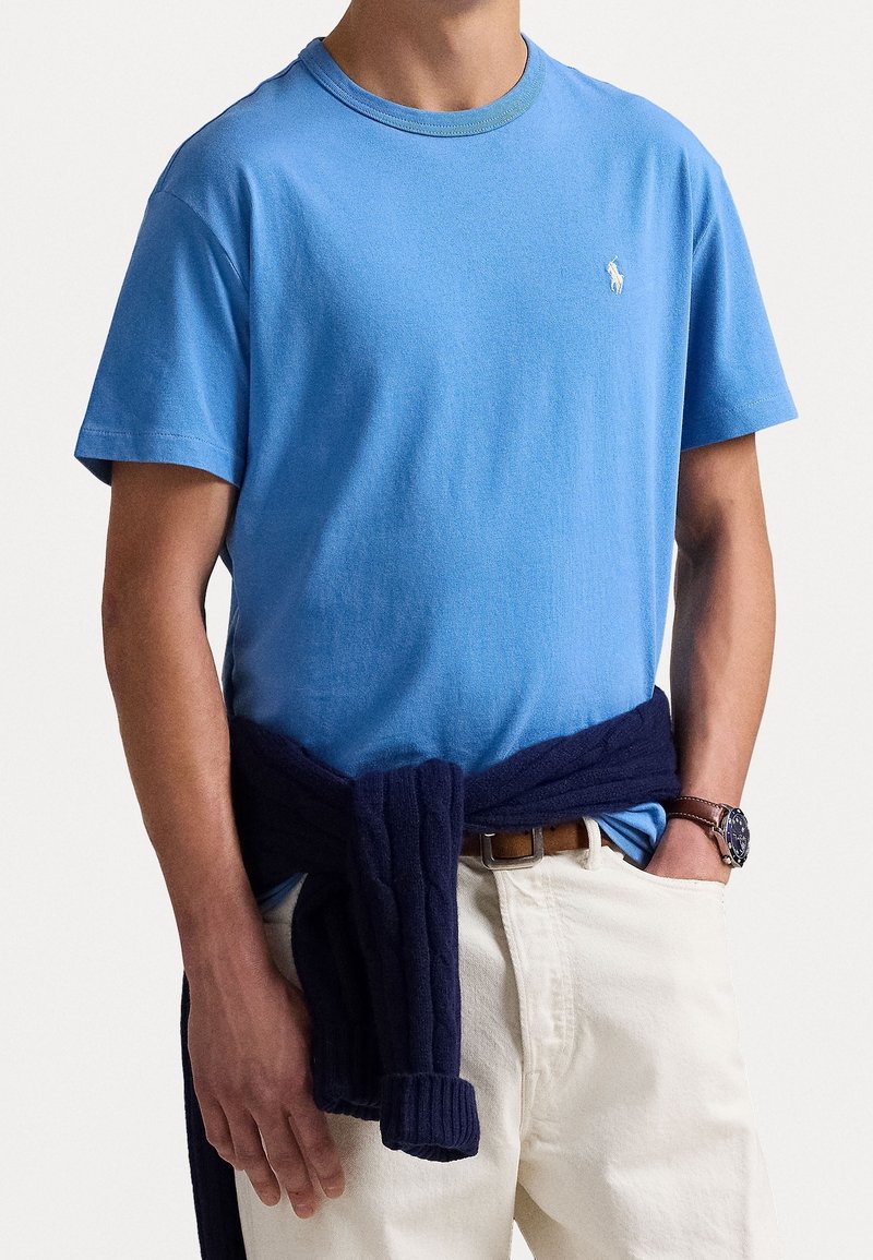 Camiseta de algodón azul con cuello redondo, mangas cortas y un pequeño logo. Suéter de punto navy atado alrededor de la cintura, combinado con pantalones beige.