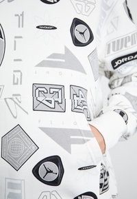 Balta kapuce ar grafisku apdruku pelēkā un melnā krāsā, attēlojot dažādus logotipus un ģeometriskas formas. Iekļauta priekšējā kabata.
