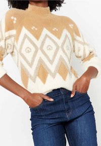 Pull en tricot crème et fauve avec un col montant, présentant un motif géométrique en forme de diamant et une finition texturée. Associé à un jean bleu foncé.