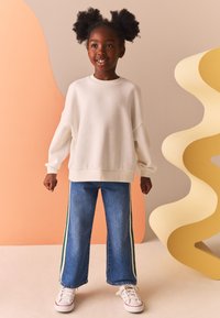 Felpa bianca oversize con spalle scese, abbinata a jeans larghi azzurri con strisce laterali verdi e sneakers bianche.