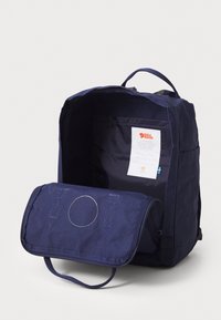 Mochila azul escura com um compartimento principal aberto, pega em pé, etiqueta costurada e aba frontal com zíper encostada.